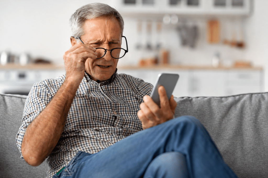 Cataract Surgery IOLs in Raleigh 1 Older man holding his glasses while struggling to read a smartphone, illustrating age-related near vision changes that may lead patients to consider presbyopic lens exchange.